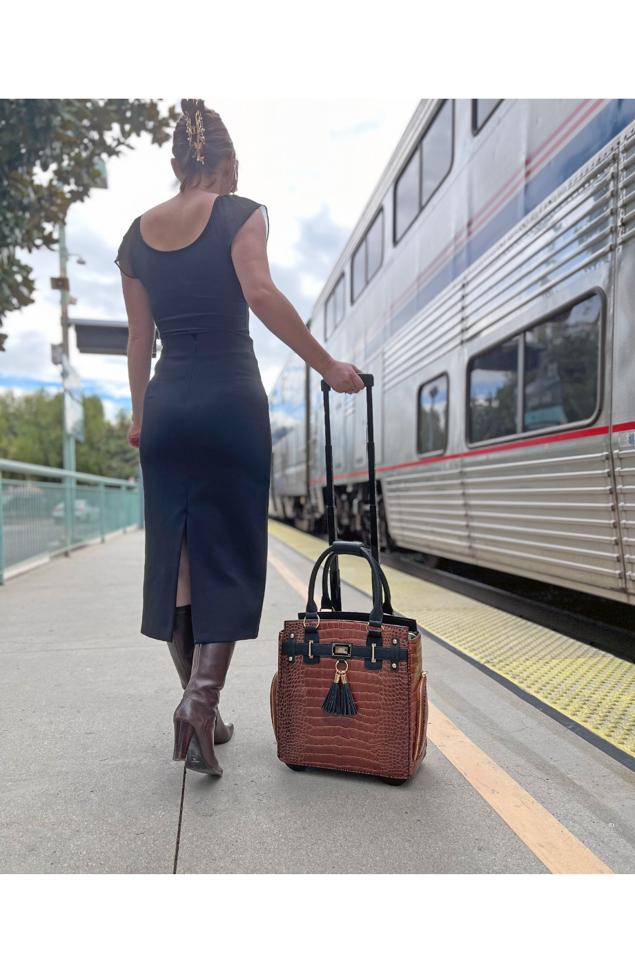 A business woman is ready to board a train on her daily commute to work. She does not have to carry any heavy bags with her because she has a cute rolling bag to hold all her work supplies. She pairs her bag with a navy maxi dress and brown boots.