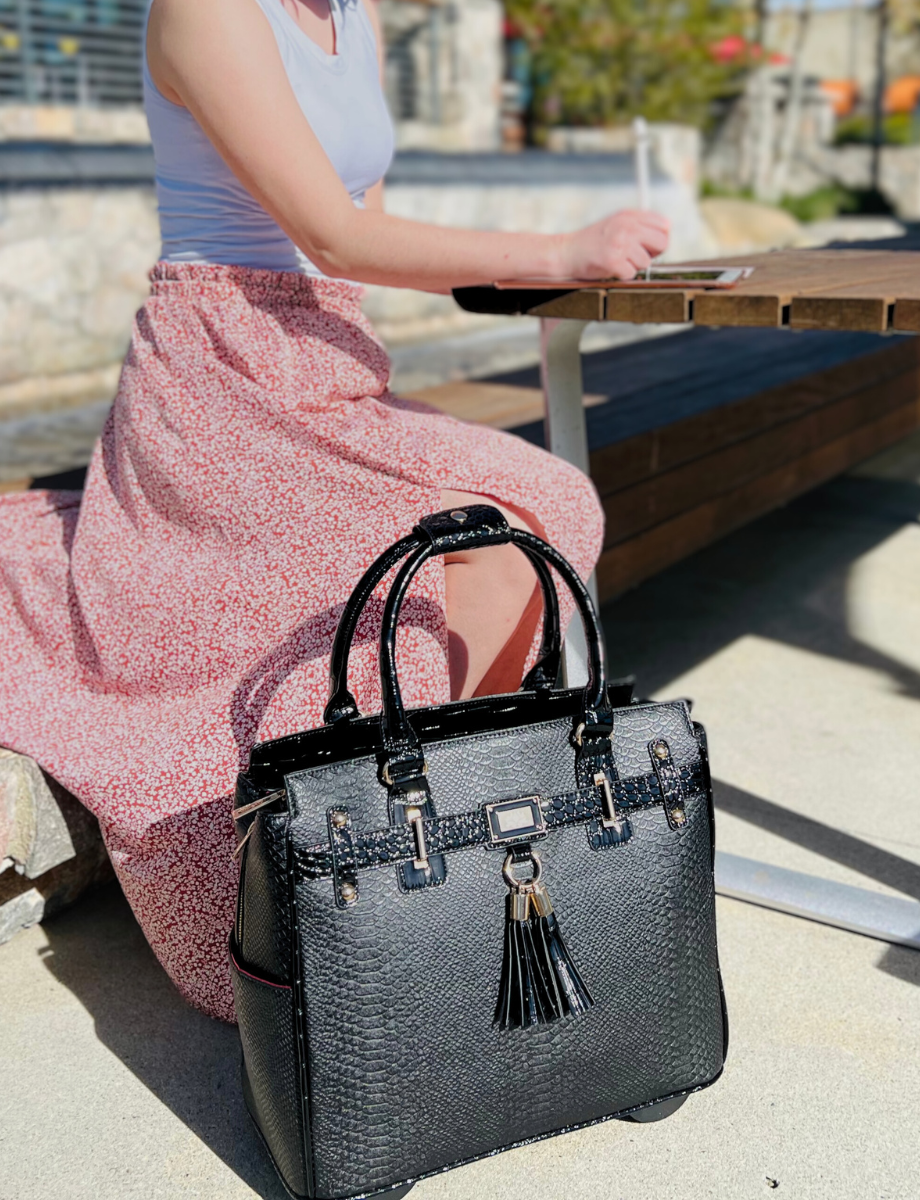 A woman is working with her stylish laptop bag 16 inch beside her, the perfect business travel bag. She can work from anywhere with her faux leather rolling bag made with vegan PU leather. She sits at a table at the park working on her ipad in a pink flower pattern flowing skirt. This versatile bag makes an excellent birthday gift ideas for mom from son. A comfort gift for her, like shoes for women, is ideal for self care gifts for her to take the weight off her shoulders on her daily commute.