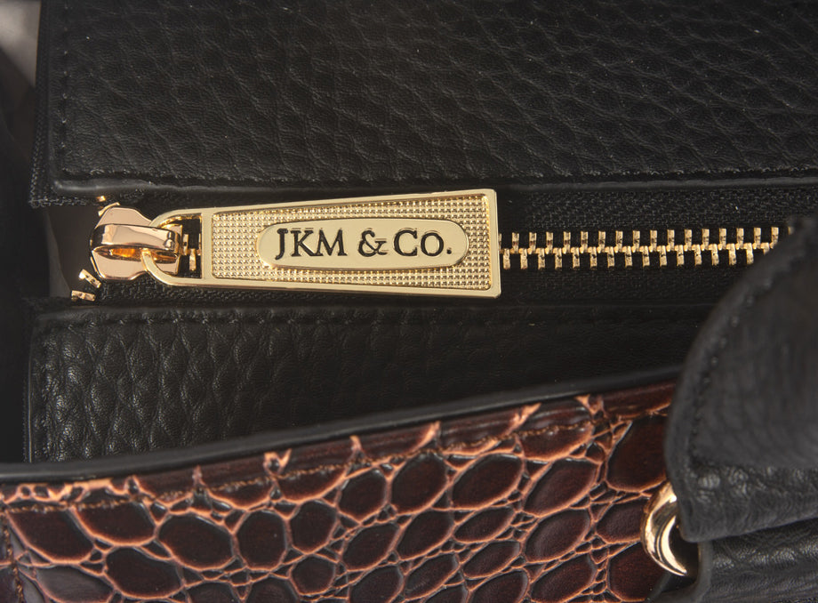 Up close image of the bag showing sleek black pebble texture fabric mixed with the rich brown alligator embossed fabric. The bag has gold metal accents including this branded YKK zipper that safely secures all your belongings in the main compartment.